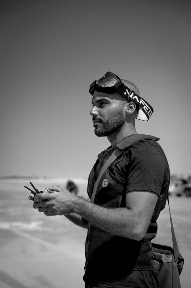 Black and white photo of a videographer with goggles holding a drone controller on the beach